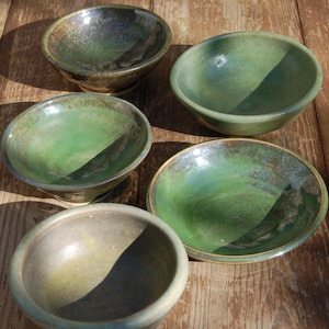 May include: Five handmade ceramic bowls in shades of green and brown. The bowls have a rustic appearance with a glossy finish. Each bowl is a unique piece, ideal for serving food or as decorative items. The bowls are of varying sizes.