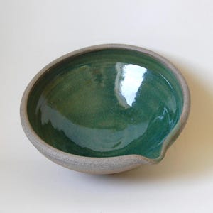 May include: A handcrafted ceramic bowl with a deep green interior and a light grey rim. The bowl has an irregular shape with a small spout on one side. The bowl is likely used for serving or as a decorative item.