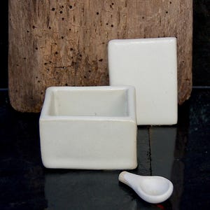 May include: A white ceramic salt cellar set. It includes a small rectangular dish, a square lid, and a spoon. The set is on a dark surface with weathered wood in the background. The salt cellar is a small, square container.