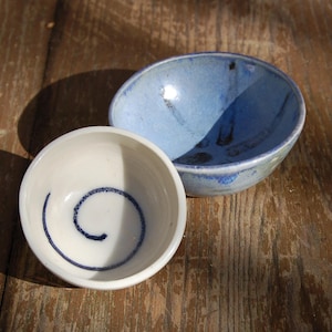 May include: Two ceramic bowls. One is white with a blue spiral design, the other is blue with darker blue accents. The bowls are on a wooden surface. The bowls are likely for serving food or as decorative items.