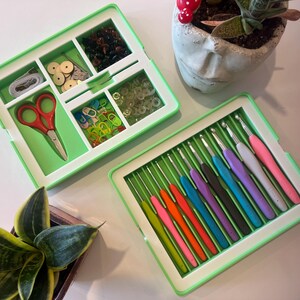 May include: Two open green craft storage boxes. One box contains sewing supplies, including red scissors, wooden buttons, and small beads. The other box holds a set of colourful crochet hooks. A small succulent plant in a face-shaped pot is also visible.