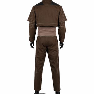 Puede incluir: Una chaqueta marr&oacute;n de manga larga con cuello alto y una capa superpuesta corta, combinada con pantalones marrones a juego. La chaqueta presenta costuras blancas en contraste a lo largo de las mangas y el dobladillo, con una banda marr&oacute;n claro en la cintura.