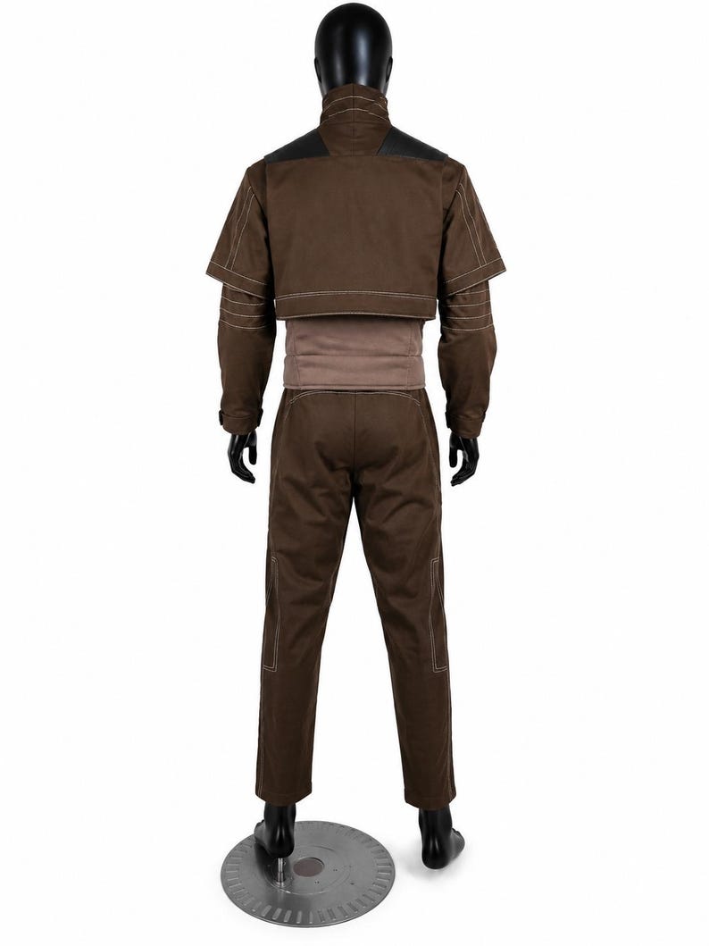 Puede incluir: Una chaqueta marr&oacute;n de manga larga con cuello alto y una capa superpuesta corta, combinada con pantalones marrones a juego. La chaqueta presenta costuras blancas en contraste a lo largo de las mangas y el dobladillo, con una banda marr&oacute;n claro en la cintura.