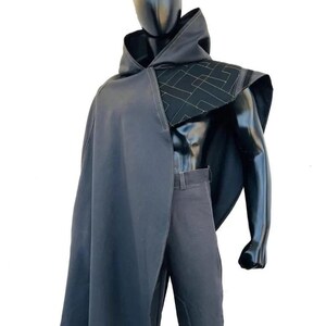 May include: A dark gray hooded cloak featuring a structured shoulder panel with a geometric gold-stitched pattern, worn over matching dark gray trousers. The cloak drapes over one shoulder, showcasing clean lines and a minimalist aesthetic design.