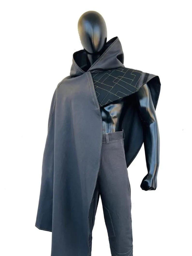 May include: A dark gray hooded cloak featuring a structured shoulder panel with a geometric gold-stitched pattern, worn over matching dark gray trousers. The cloak drapes over one shoulder, showcasing clean lines and a minimalist aesthetic design.