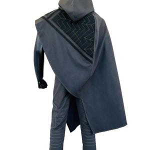 May include: A dark gray hooded cloak featuring a black triangular panel with a gold geometric zigzag pattern on the back. The cloak is worn over matching dark gray trousers with horizontal stitched detailing on the legs, presented on a mannequin.