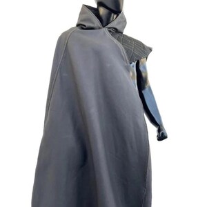 May include: A black hooded cloak featuring a structured shoulder panel with a geometric quilted pattern and visible light-colored stitching. The garment drapes over the torso, showcasing a smooth fabric texture and a high collar design.