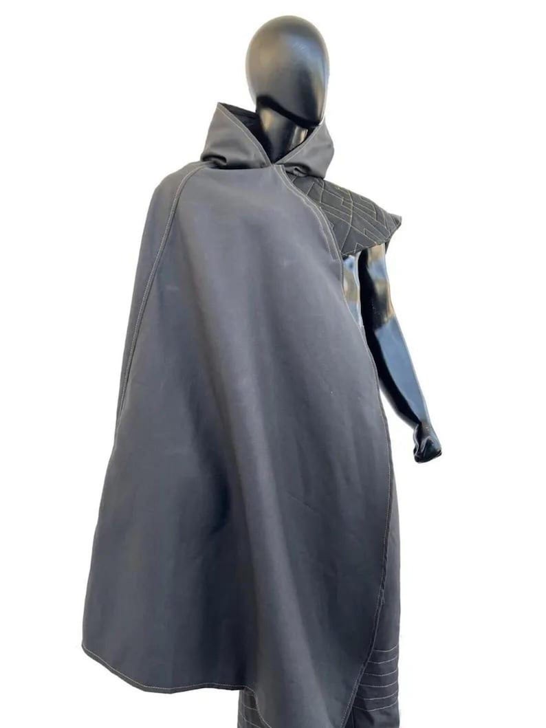May include: A black hooded cloak featuring a structured shoulder panel with a geometric quilted pattern and visible light-colored stitching. The garment drapes over the torso, showcasing a smooth fabric texture and a high collar design.