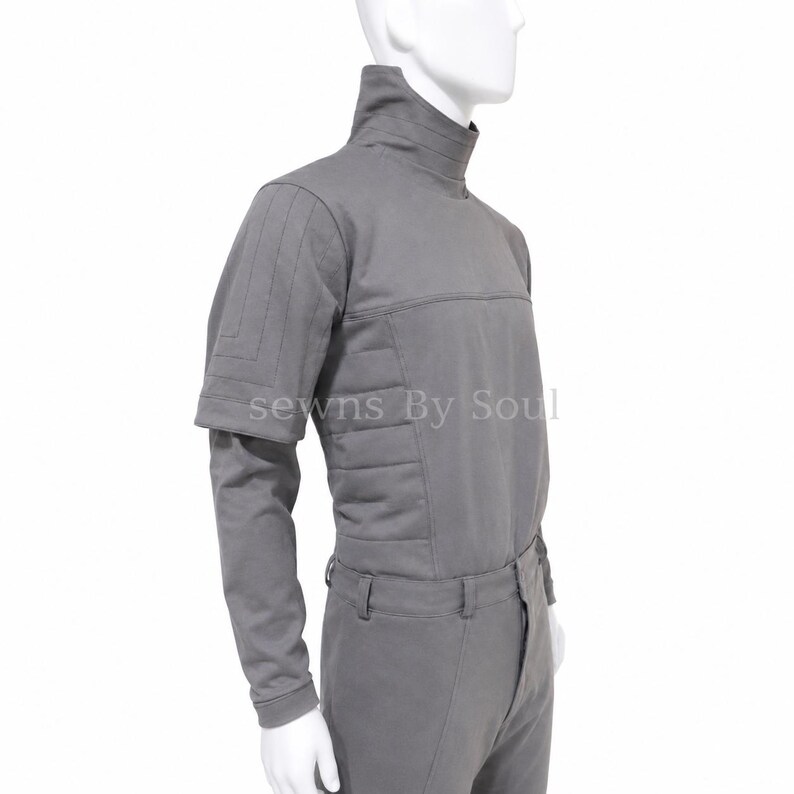 K&ouml;nnte beinhalten: Ein graues Langarmshirt mit hohem Kragen und strukturierten Eins&auml;tzen an den Schultern und Seiten. Das Kleidungsst&uuml;ck weist ein horizontal gestepptes Design an den Ober&auml;rmeln und am Rumpf auf. Der Text "sewns By Soul" ist auf der Brust sichtbar.