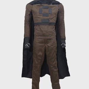 May include: A brown long-sleeved shirt and matching trousers set featuring a black cape, a high neck collar, and dark rectangular patches on the chest. The sleeves display white geometric stitching patterns, and the garment is displayed on a mannequin.