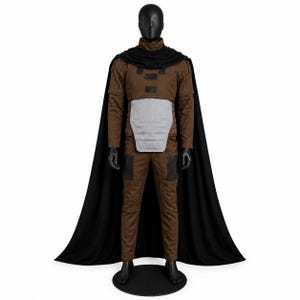 May include: A brown long-sleeved jumpsuit with a high collar, featuring a light gray abdominal armor plate and black rectangular accents on the chest and thighs. A long black cape drapes from the shoulders over the brown jumpsuit ensemble.
