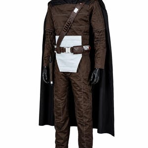 Puede incluir: Un mono marr&oacute;n de manga larga con detalles de costuras blancas, combinado con una capa negra, un panel de cintura rectangular blanco y un cintur&oacute;n utilitario marr&oacute;n con hebilla plateada, una peque&ntilde;a bolsa y una funda para un accesorio.