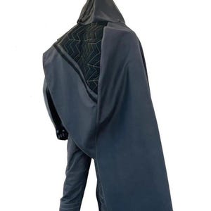 May include: A dark gray hooded cloak featuring a black quilted panel on the upper back with a white zigzag stitch pattern. The cloak is worn over matching dark gray trousers that have horizontal stitched lines around the lower leg area.