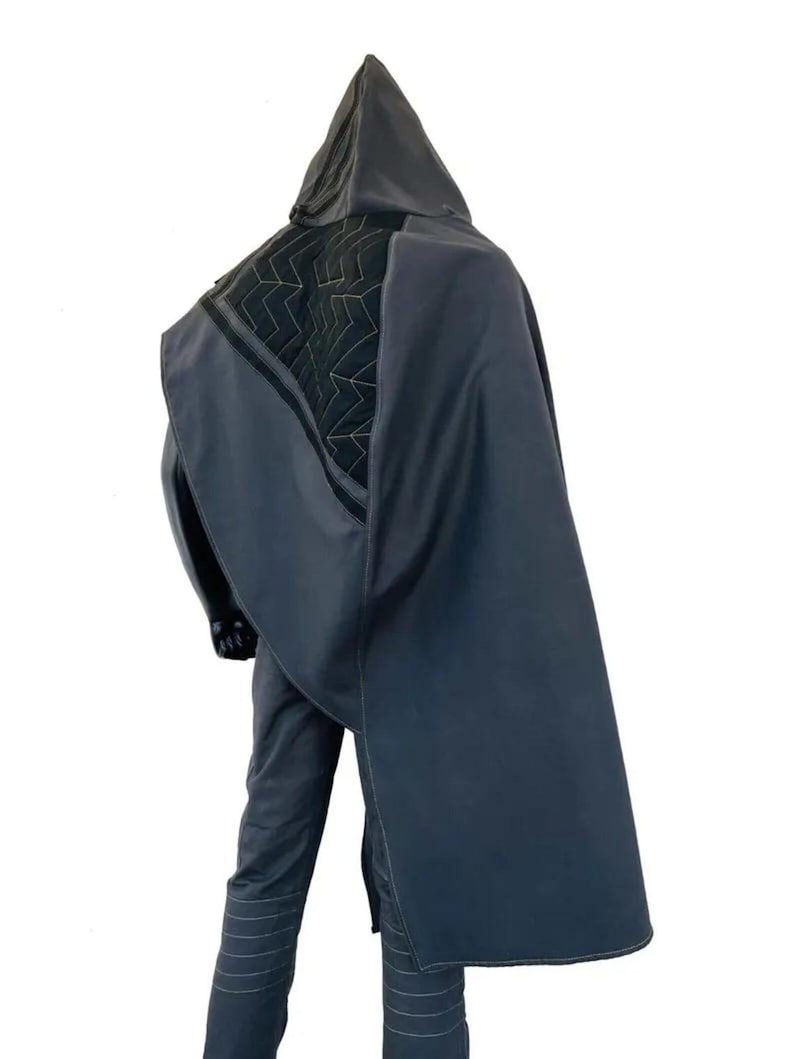 May include: A dark gray hooded cloak featuring a black quilted panel on the upper back with a white zigzag stitch pattern. The cloak is worn over matching dark gray trousers that have horizontal stitched lines around the lower leg area.