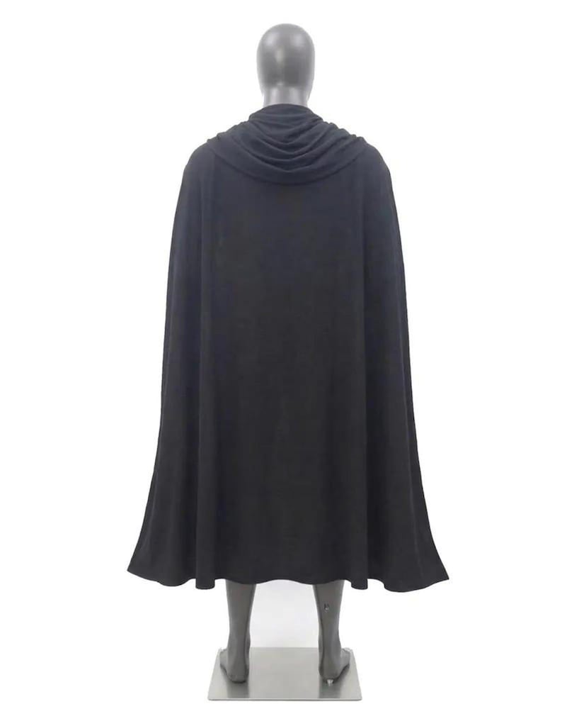 May include: A solid black floor-length cape featuring a draped cowl neckline that rests around the shoulders. The garment is displayed on a neutral-toned mannequin, showcasing a full-length silhouette with a smooth, flowing fabric texture.