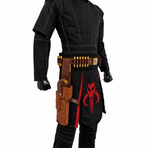 May include: A black long-sleeved tunic and pants set featuring white stitching, a brown leather belt with gold-colored ammunition loops, a matching brown leather holster, and a black apron panel displaying a large red skull-shaped emblem.