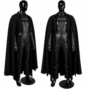 May include: A full-body black costume displayed on two mannequins, featuring a long cape, a quilted textured tunic with long sleeves, matching trousers, and a structured belt piece. The ensemble is entirely black with varied fabric textures.