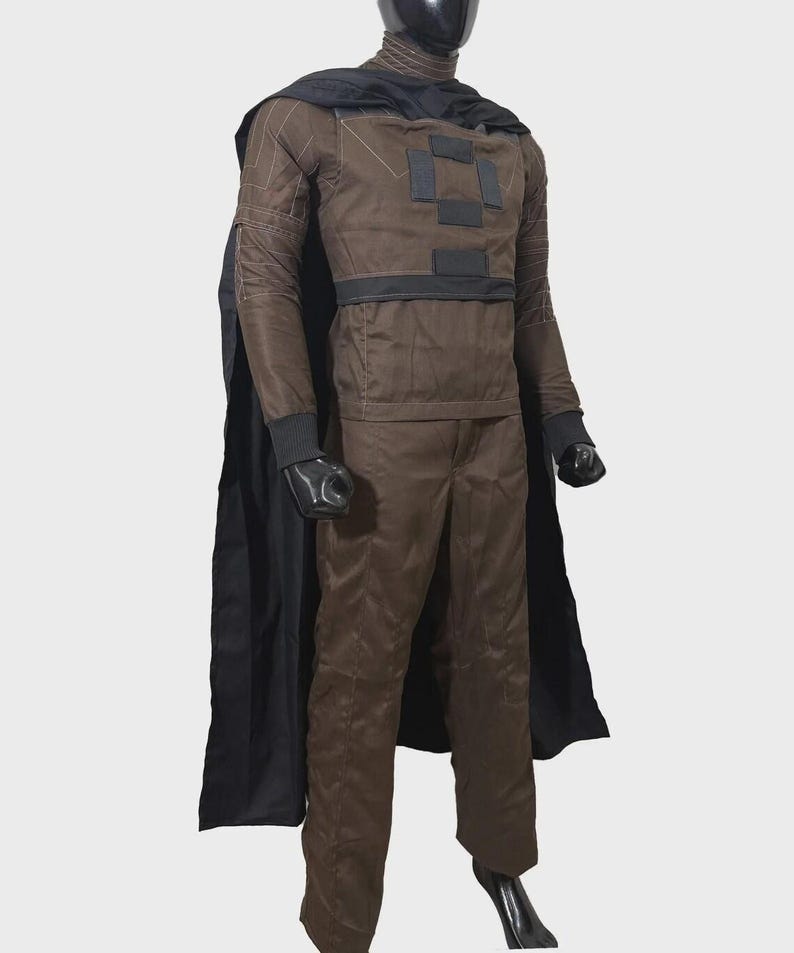 May include: A brown long-sleeved shirt and matching trousers costume set featuring a black cape, a high collar, and black rectangular patches on the chest. The sleeves have white stitched detailing, and the cuffs are ribbed in black fabric.