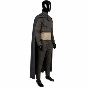 May include: A dark gray long-sleeved tunic and matching trousers set worn with a long, flowing dark gray cape. The outfit includes a wide, light tan horizontal waistband and features structured, quilted-style stitching on the upper sleeves.