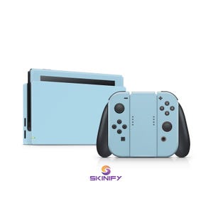 Może przedstawiać: Jasnoniebieska konsola Nintendo Switch i kontrolery. Kontrolery mają czarne akcenty i przyciski. Konsola i kontrolery są umieszczone na białym tle. Logo Skinify znajduje się na dole.
