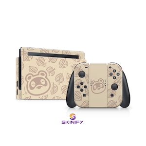Könnte beinhalten: Eine beige Nintendo Switch-Konsole und -Controller mit einem sich wiederholenden Blättermuster und einem Cartoon-Waschbärgesicht. Die Controller haben schwarze Akzente und Tasten. Das Skinify-Logo befindet sich unten.