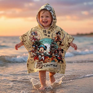 Op de afbeelding: Een beige poncho met capuchon voorzien van een piraten-thema grafische print met stripfiguren, een schip en de tekst "YO HO YO HO" en "CYNTHIA". Het ontwerp bevat diverse als piraat verklede figuren en een scheepsroer op een perkamentachtige achtergrond.