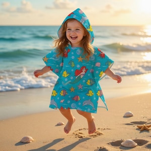 May include: A child wearing a turquoise hooded beach towel with a colorful Little Mermaid design, including Ariel, fish, and starfish. The child is jumping on a sandy beach near the ocean at sunset.