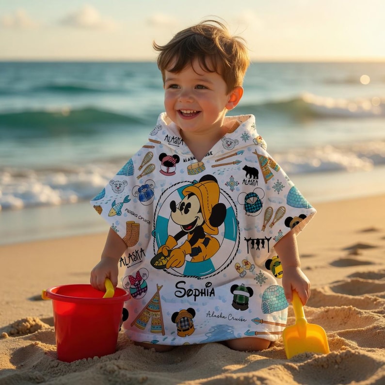 Puede incluir: Un poncho de playa con capucha blanco que presenta un gr&aacute;fico central de Mickey Mouse con impermeable amarillo dentro de un salvavidas azul. La tela tiene iconos peque&ntilde;os como osos, igl&uacute;s y tiendas, con el nombre Sophia impreso en negro.