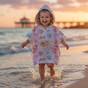 May include: A young child wearing a pink hooded beach towel with a pattern of desserts and bows, running through the water at the beach. The towel has a white and pink pattern with images of cakes, hearts, and cherries. The child is smiling.