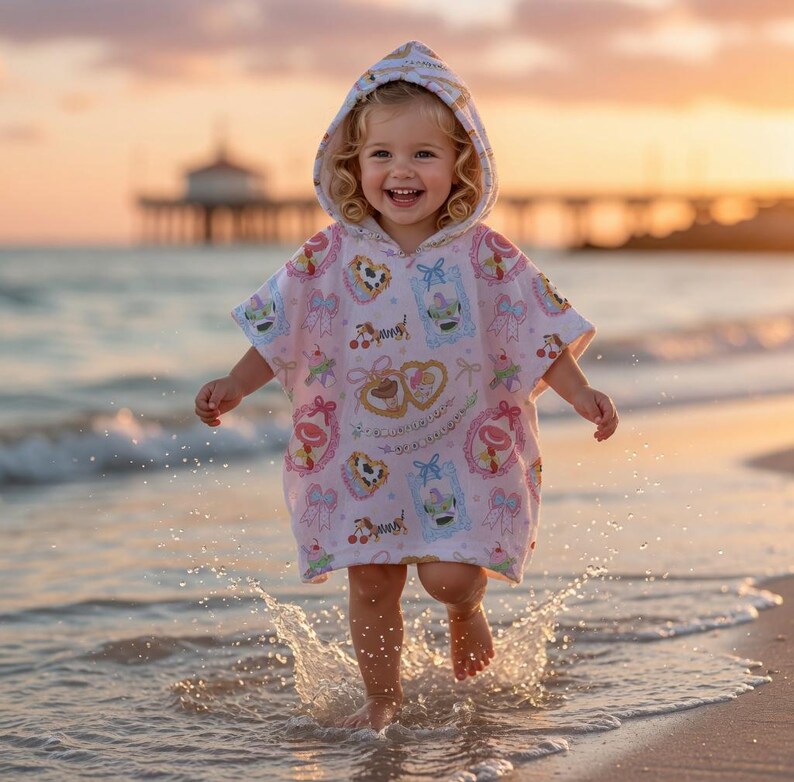 May include: A young child wearing a pink hooded beach towel with a pattern of desserts and bows, running through the water at the beach. The towel has a white and pink pattern with images of cakes, hearts, and cherries. The child is smiling.