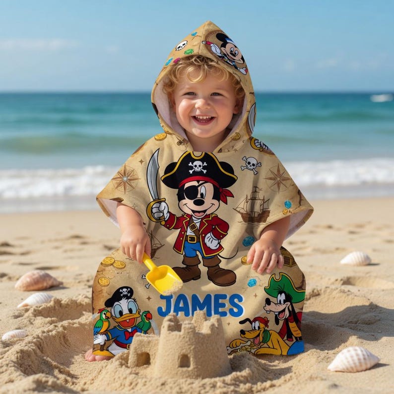 May include: A tan hooded beach towel featuring a pirate-themed Mickey Mouse design, with a pirate hat, eye patch, and sword. The towel also features Donald Duck and Goofy. The name "JAMES" is printed in blue.