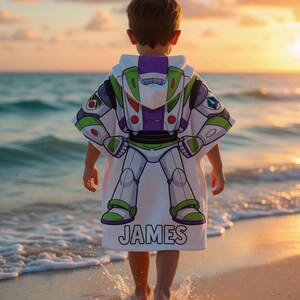 May include: A white hooded beach towel with a Buzz Lightyear design, featuring green, purple, and white details. The name "JAMES" is printed in black at the bottom. A child wears the towel while standing on a sandy beach, near the sea.