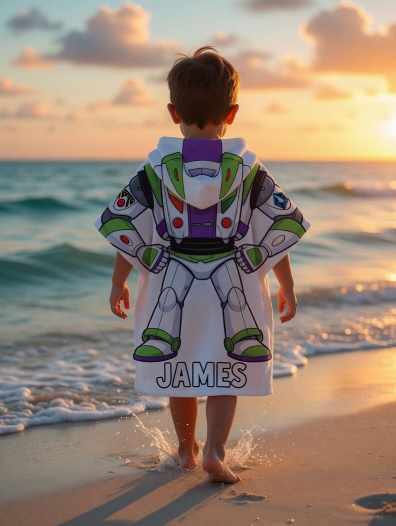 May include: A white hooded beach towel with a Buzz Lightyear design, featuring green, purple, and white details. The name "JAMES" is printed in black at the bottom. A child wears the towel while standing on a sandy beach, near the sea.