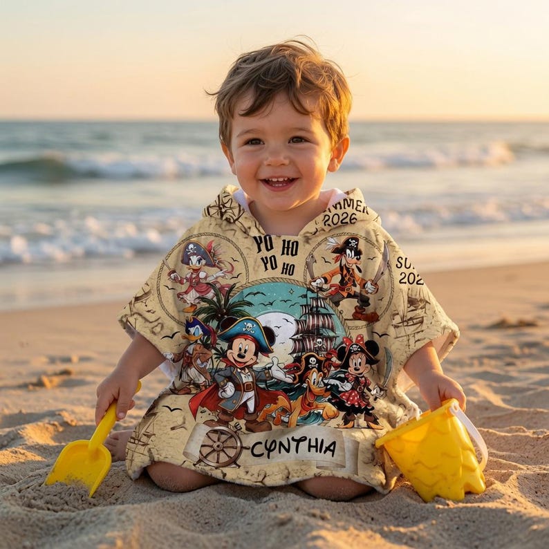 Op de afbeelding: Een strandponcho met capuchon op een beige achtergrond met een piraten-grafische print van stripfiguren, een schip en de tekst "YO HO YO HO" en "CYNTHIA". Het ontwerp bevat diverse nautische elementen en figuren in piratenkleding.
