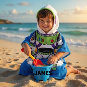 May include: A child wearing a blue and white hooded beach towel with a Buzz Lightyear design, playing with a red and yellow shovel and a blue bucket on a sandy beach. The bucket has the name "JAMES" printed on it.