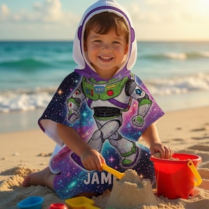 May include: A child wearing a hooded beach towel featuring a Buzz Lightyear design. The towel has a white hood with purple trim and a colorful graphic of the character against a galaxy background. The child is playing with sand toys on a sandy beach.