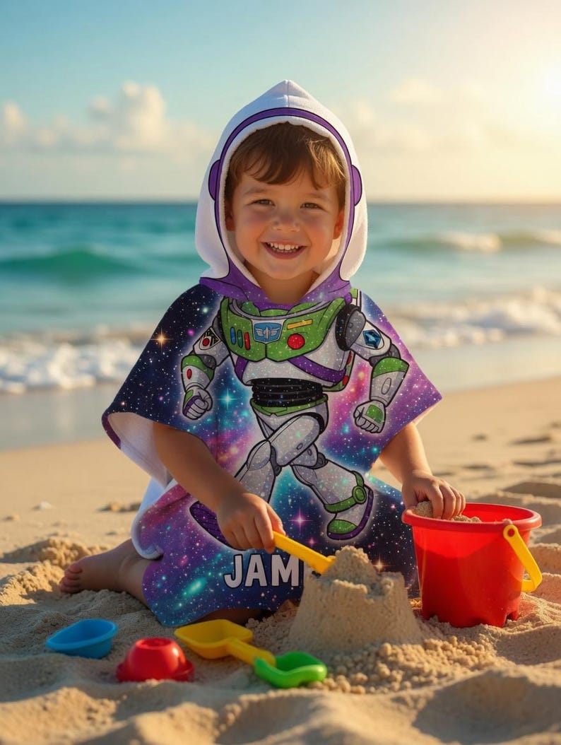 May include: A child wearing a hooded beach towel featuring a Buzz Lightyear design. The towel has a white hood with purple trim and a colorful graphic of the character against a galaxy background. The child is playing with sand toys on a sandy beach.