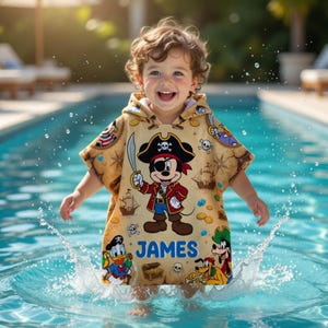May include: A child wearing a tan pirate-themed hooded towel with Mickey Mouse, Donald Duck, and Pluto graphics. The towel features a pirate map design, the name "JAMES" in blue, and a pirate ship illustration. The child is in a pool.