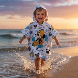 Puede incluir: Un poncho de ba&ntilde;o blanco con capucha que presenta un gr&aacute;fico central de Mickey Mouse con un sombrero amarillo y un salvavidas azul. La tela est&aacute; decorada con iconos tem&aacute;ticos de Alaska y el nombre Sophia impreso en letra cursiva negra.