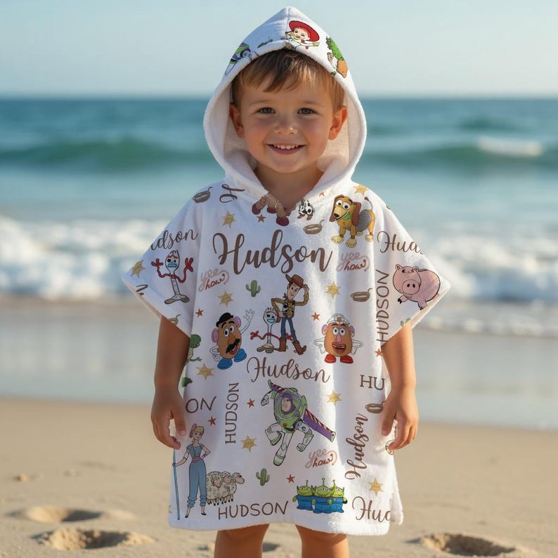 May include: A white hooded poncho towel featuring a repeating pattern of Toy Story characters including Woody, Buzz Lightyear, and Mr. Potato Head, interspersed with gold stars, cacti, and the name Hudson written in a cursive brown script font.