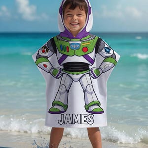 May include: A white hooded beach towel with a Buzz Lightyear design, featuring green, purple, and white details. The towel displays a graphic of the character's spacesuit and the name "JAMES". The child is standing on a beach with the sea in the background.
