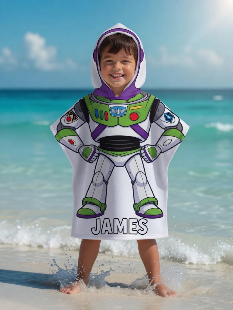May include: A white hooded beach towel with a Buzz Lightyear design, featuring green, purple, and white details. The towel displays a graphic of the character's spacesuit and the name "JAMES". The child is standing on a beach with the sea in the background.
