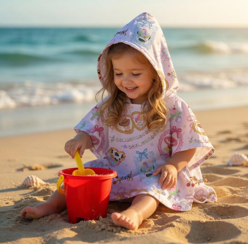 May include: A child wearing a pink and white hooded beach poncho with a pattern of hearts, bows, and other decorative elements. The child is playing with a red bucket and yellow shovel on a sandy beach near the ocean.