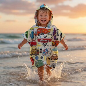 May include: A child wearing a hooded beach towel featuring a car and truck design, with the name "Henry". The towel is yellow and decorated with various vehicle illustrations. The child is at the beach.