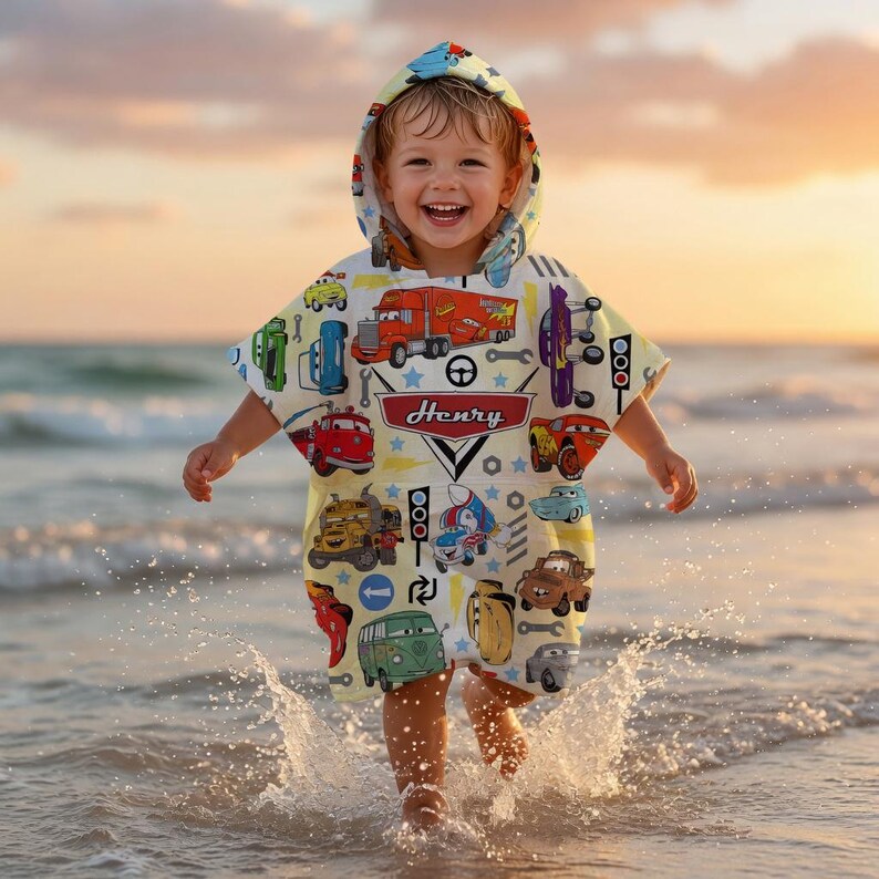 May include: A child wearing a hooded beach towel featuring a car and truck design, with the name "Henry". The towel is yellow and decorated with various vehicle illustrations. The child is at the beach.