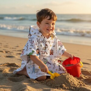 May include: A white hooded towel poncho featuring a repeating pattern of colourful cartoon characters from the Toy Story franchise, including Woody, Buzz Lightyear, and Jessie, interspersed with the name Hudson in a brown cursive script font.