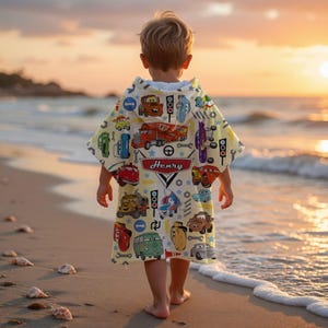 May include: A child wearing a yellow hooded beach towel with a colourful car and truck pattern, and the name "Henry" printed on it. The child is walking on a sandy beach near the ocean at sunset.