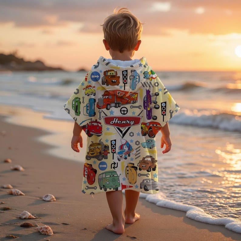 May include: A child wearing a yellow hooded beach towel with a colourful car and truck pattern, and the name "Henry" printed on it. The child is walking on a sandy beach near the ocean at sunset.
