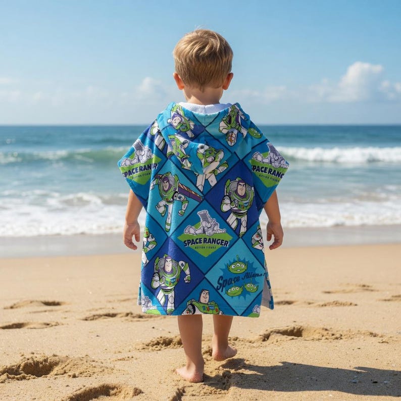 Może przedstawiać: Niebiesko-zielony ręcznik plażowy z kapturem z postacią Buzz Lightyear z Toy Story. Ręcznik ma wz&oacute;r w romby z wizerunkami Buzza Lightyeara oraz napisami "Space Ranger" i "Space Aliens". Dziecko stoi na piaszczystej plaży.