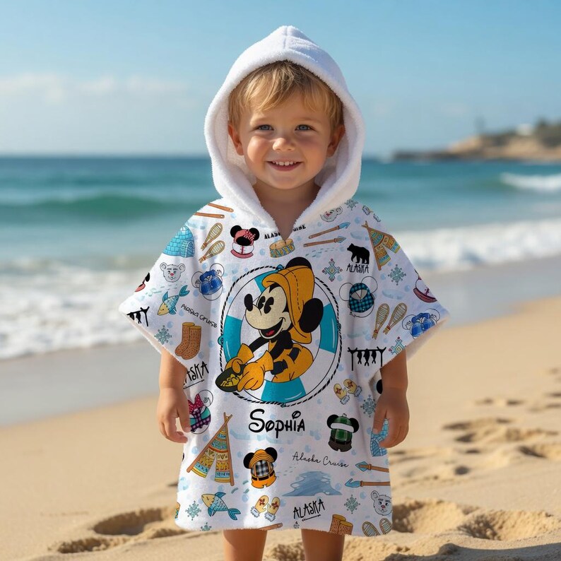 Puede incluir: Un poncho de toalla blanco con capucha que presenta un gr&aacute;fico central de Mickey Mouse con sombrero y abrigo de lluvia amarillos. La tela tiene ilustraciones de peces, botas, tipis y el texto "Sophia" y "Alaska Cruise" en color negro.